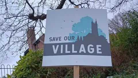 A cardboard sign on a wooden stick reading 'Save our village'  placed near bushes on the side of a road. Behind the sign bushes and a red brick house can be seen. 