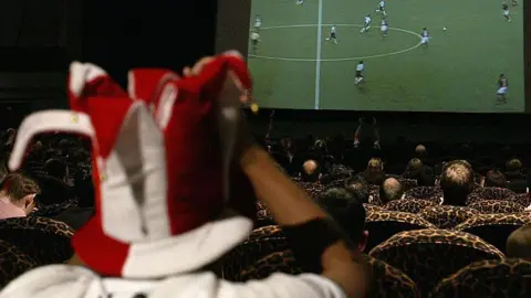 Getty Images Cinema attendees sitting in patterned theatre seats watching a football match on a large screen, with a fan in a red and white jester hat in the foreground.