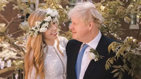 Rebecca Fulton/Downing Street/PA Wire Boris Johnson standing next to his wife Carrie with his arm around her. He is wearing a black suit, white shirt and blue tie. Carrie is wearing a lace wedding dress with intricate detailing down the middle and on the sleeves. She is also wearing a white rose flower crown and smiling at her husband. They are standing on an outdoor staircase next to a bush with pink flowers on it.