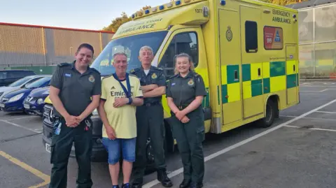 A man wearing a yellow Arsenal football shirt stood with three paramedics, with an ambulance behind them. 