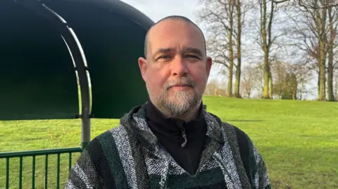Rob Chapman is standing outside in a park with trees in the background. He is wearing a grey and black top and has a grey beard.