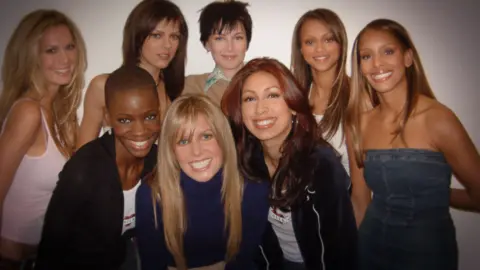 Netflix Ebony Haith (bottom left) smiles at a camera alongside fellow cycle one contestants. She is wearing a black cardigan over a white top and has a shaved head. 