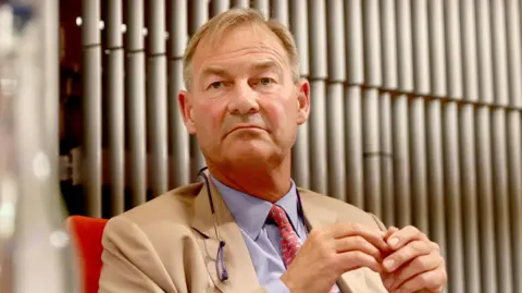 PA Media Rupert Lowe with short blond hair wearing a cream jacket, light purple shirt and red patterned tie.