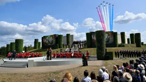 PA Media The VJ Day service held near the central memorial at the National Memorial Arboretum. People are watching on as military personnel stand to attention. The Red Arrows, leaving red, white and blue smoke trails, can be seen rising up from behind the memorial.