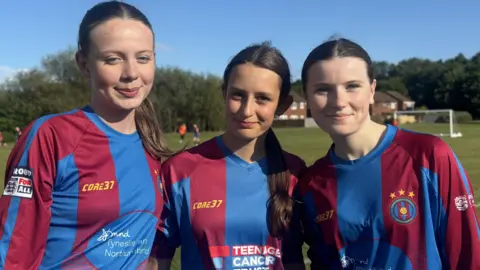 Three footballers stand together wearing blue and red striped football tops. They all have dark hair which is tied back and they are standing at the edge of a football pitch