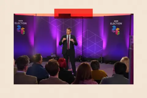 Plaid Cymru leader Rhun ap Iorwerth at a BBC Wales Ask the Leaders debate in Haverfordwest, Pembrokeshire. He is wearing a dark suit and a white shirt with a purple tie and stands in front of an audience, whose heads are visible from the rear. The words "BBC ELECTION 26" appear in two places on a screen behind him.