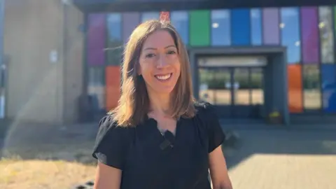Sarah has a black short sleeve top and is smiling in front of a colourful building on a sunny day.