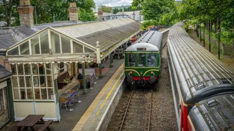 John Askwith And John Dinsdale A train pulling into Bishop Auckland West station. A man sits at a table on the covered platform.
