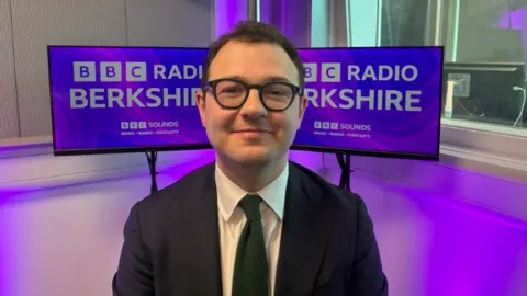 Windsor MP Jack Rankin smiling at the camera is wearing a suit, green tie and dark framed glasses.
