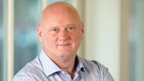 FICO A bald man smiles at the camera. He wears a blue checked shirt. Blurred windows can be seen behind him.
