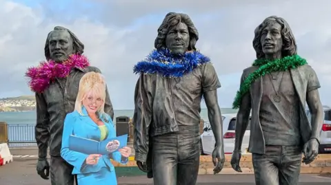 A life-size cutout of Dolly Parton reading with the Beegees statue. Each member of the band has tinsel around their necks as scarves.