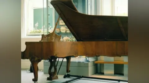 Sunderland Pianoforte Society has ornate legs and is in light walnut colour. Some ceramics are stored in glass cupboard behind it. 