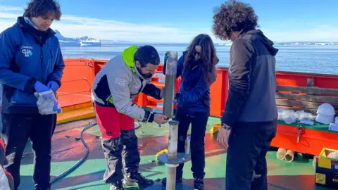 Elisenda Balleste Four researchers are on board the deck of a ship removing a large tube of sediment from a core drilling machine. There is bright sunshine, but the scientists are all wearing heavy coats and the icy scenery of the Antarctic Peninsula is visible in the background 