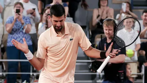 Novak Djokovic, wearing a peach polo shirt, dances with his racquet in his left hand