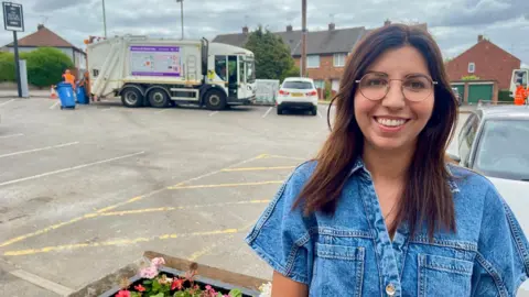 A woman with long hair and glasses is stood in a car park. There is a bin lorry behind her.