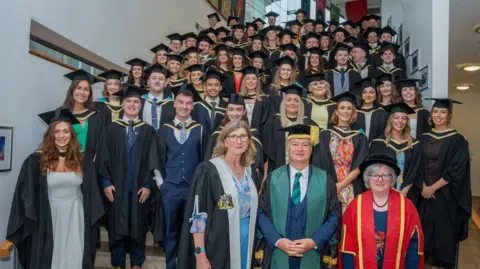 a group of 69 medical graduates stand on the stairs of Derry's millenium forum following their gradution. the men and women are wearing formal dress and mortar boards and gons