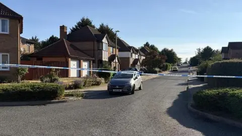 Amy Holmes/BBC A street in a housing estate with houses on both sides. There is a line of blue and white police tape stretched across the road and a car immediately behind it on the left.