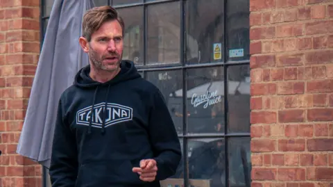 Supplied A man stand outside a red-brick building with large arched windows and black metal frames. The man is wearing a dark hoodie with a logo points while speaking.