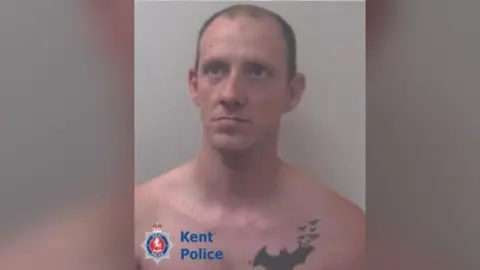 Sean Boyle - a close up police mugshot of a man naked from the top up with a large bat tattoo on his left breast. He is looking seriously to the camera's right