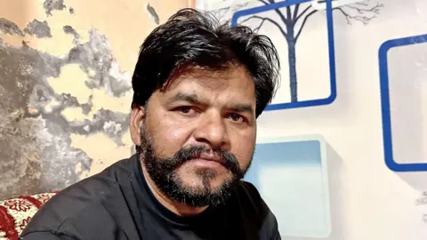 BBC Gurpreet Singh, an Indian man with black hair and black hair cut in the back and sides, but long on top, wears a black shirt while sitting in a room with a mural on one wall that includes trees and geometric shapes, while on the other wall, the plaster collapses.