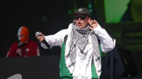Mo Chara of Kneecap holds a hand to his ear during the group's performance at Glastonbury. He is wearing a black baseball cap, sunglasses, white jacket and Palestinian keffiyeh. Another band member wearing a tricolour balaclava is in the background 