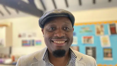 A man with a short black beard. He is wearing a hat. He is smiling at the camera. He is wearing a light jacket and striped shirt.