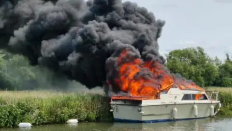 A white boat on fire with black smoking billowing from it. Beyond it is a river bank, a field, and trees in the background. 