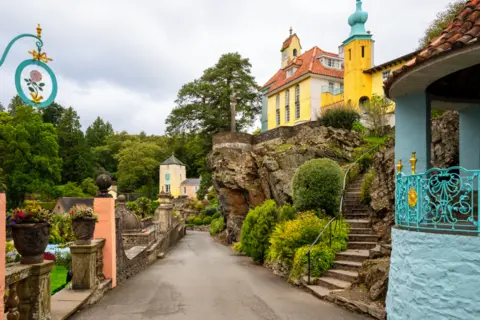 Getty Images Buildings in Portmeirion. A road runs in the middle of the photo. To the right is a turquoise mini pavillion. Steps lead up to a yellow house with a turquoise turret and terracotta tiled roof in the Italian style. It stands on an outcrop of rock. To the left are the walls of gardens including a couple of peach painted pedestal supports. A couple of buildings painted yellow and light orange are in the distance.