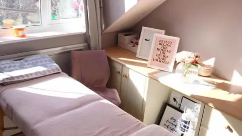 Lucy Rebecca Photography A small bedroom with a flat bed with pink cover on it. There is a pink chair next to it and a wooden side board, with flowers and photos on it.
