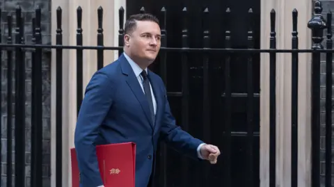 Getty Images Health secretary Wes Streeting walking on Downing Street. He is wearing a blue shirt and tie. He is holding a red binder under his right arm.