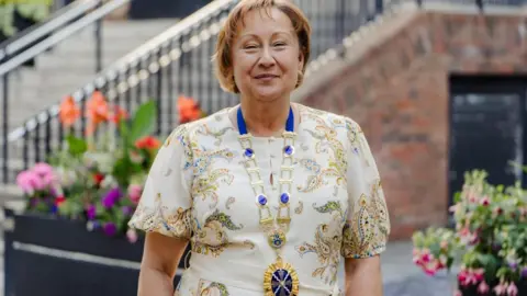 Rachel Nolan is smiling at the camera standing outdoors in front of flower beds and a staircase. She is wearing a patterned cream dress and a presidential gold and blue chain around her neck.