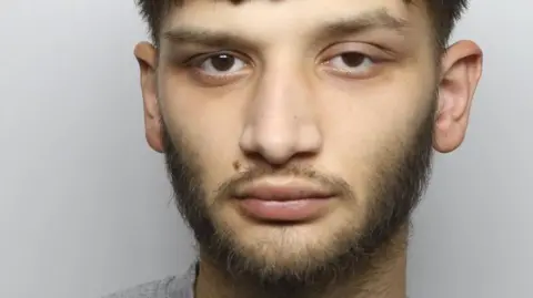 A police mugshot of Peter Krok. He has a beard and dark hair, and is wearing a grey top 