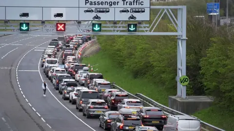 PA Media Passengers queue to use the Channel Tunnel in Folkestone, during the Easter getaway on 3 April 2026. Long queues of cars and vans wait on a multi-lane road approaching the terminal. An overhead sign marked "Passenger Traffic" displays lane information for cars, coaches and motorcycles, with illuminated green arrows and a red cross above the lanes.