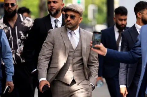 A man in a beige three-piece suit and a beige cap walks outside, surrounded by other men in suits. The man is wearing sunglasses and he has a neatly trimmed black beard.