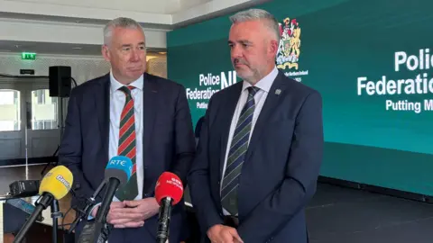 Two men with short grey hair, stand in front of three microphones. The mics are yellow, blue and red. The man on the left wears a navy suit and white shirt with a red and green striped tie. The man on the right also wears a navy suit and white shirt with a green and navy striped tie. Behind them is a small stage with a large green sign which says 'Police Federation NI. Putting Members First."