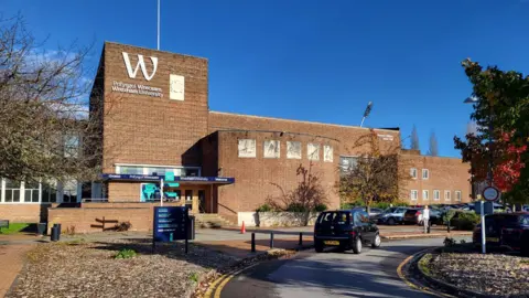 Frontage of a light brown brick university building with trees either side, set against a very blue sky