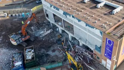 A two-storey building is being demolished with orange construction machines on a building site, surrounded by planks of wood and other items.