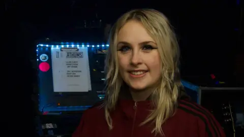 John Fairhall/BBC Bridget has blonde shoulder-length hair. She wears a dark red tracksuit top which is zipped up. She has dark eyeliner and is smiling at the camera. She stands in front of a music mixing desk.
