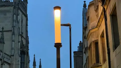 Steve Hubbard/BBC A lamp lit up against the evening sky. It is tall and thin and there are ornate university buildings on either side