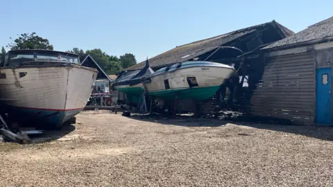 Owen Sennitt/BBC A white-painted Broads cruiser and two other boats with blue and white hulls that have been badly damaged in a fire can be seen next to a fire-damaged wooden boat shed