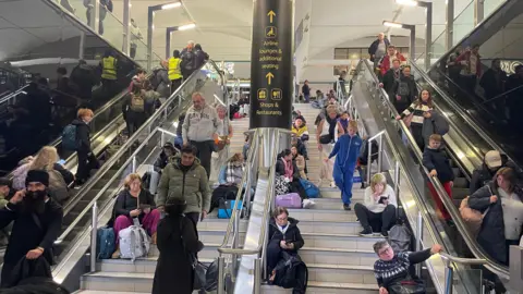 People sitting along stairs and travelling on escalators in both directions during flight delays and cancellation at Gatwick on 28 December