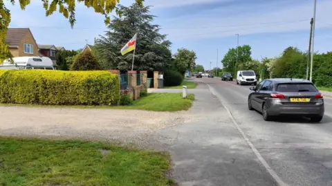 Local Democracy Reporting Service An entry to a gravel road, with grass on either side of the gravel. On the right is a single-carriageway road with cars and vans travelling in both directions. On the far side of the gravel road is a hedge with house-roofs and a campervan roof above