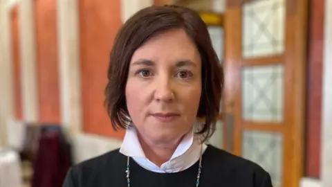 A woman with bob length dark hair stares into the camera. She is wearing a white shirt with the collar up with a dark jumper over it. She has a long silver chain around her neck. In the background is a set of double doors and a some chairs and a table