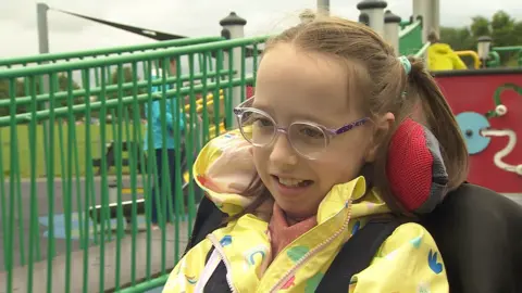 A picture of Sophie's daughter Millie. She is in a wheelchair and wearing a yellow raincoat, with purple-pink glasses. She has blonde hair.