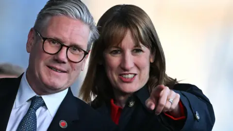 Prime Minister Keir Starmer stands next to Rachel Reeves, who is pointing to something ahead. Starmer is bespectacled and wearing a suit with a poppy attached to his jacket. Reeves is wearing a jacket with what looks like a red shirt underneath.