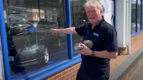 Dusty Chelmsford A man standing in front of a shop window with a very large crack in it He is holding two fist-sized stones