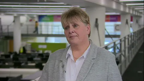 A woman with short blonde hair stands on a gallery overlooking a space below with desk and computers. She wears a white blouse with a grey jacket over the top and has no expression as she looks past the camera.