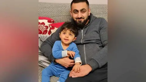 PA Media Eisa and his father Safdar smiling at the camera and sitting on a sofa. Eisa is in a blue baby grow and Safdar has dark hair, eyes and a beard. He is wearing black trousers and a grey zipped hoodie.