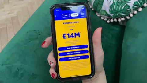 BBC A woman's hand holding a phone with the lottery app on