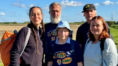 Three women, including Cecile Roberts, and two men, wearing rucksacks, and some with blue "Walk of Kindness" T-shirts. pictured in a field on their charity walk. There is a blue sky and white clouds behind them.